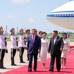 Xi Jinping and Kim Jong Un walking on a red carpet at an airfield with an honor guard and a large airplane in the background.