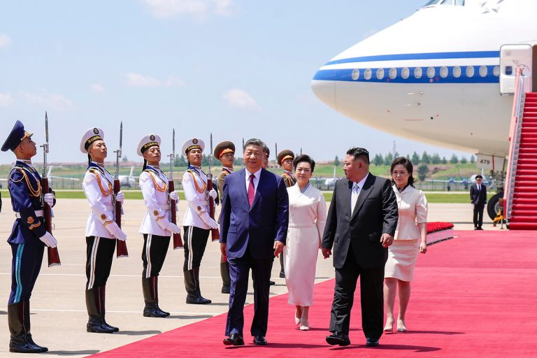 Xi Jinping and Kim Jong Un walking on a red carpet at an airfield with an honor guard and a large airplane in the background.