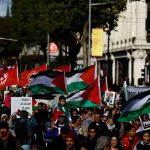 A large crowd of protestors marching in a street carrying Palestinian flags and protest signs.