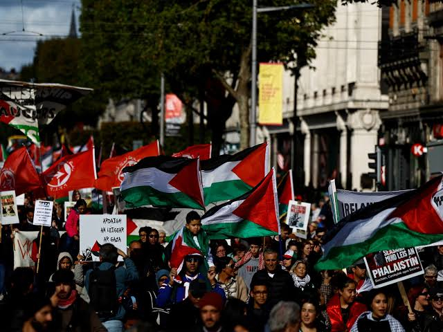 A large crowd of protestors marching in a street carrying Palestinian flags and protest signs.