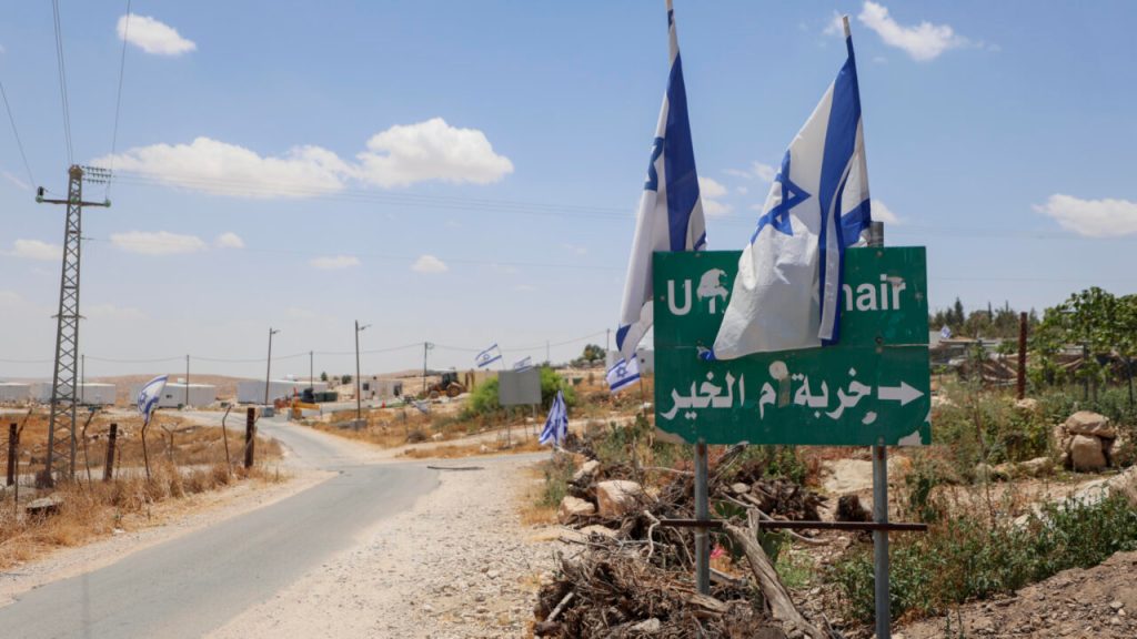 A road sign pointing toward a settlement with Israeli flags flying in the background.