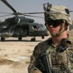A US soldier in combat gear standing in front of a military helicopter in a desert environment.