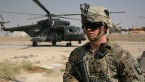 A US soldier in combat gear standing in front of a military helicopter in a desert environment.