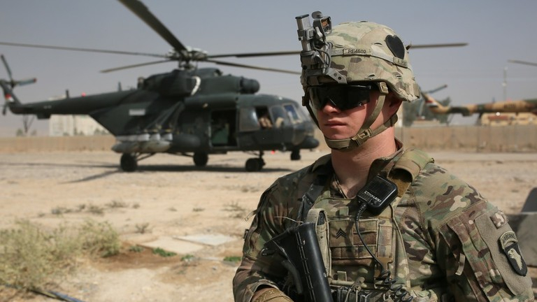 A US soldier in combat gear standing in front of a military helicopter in a desert environment.