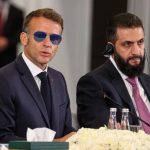 French President Emmanuel Macron wearing sunglasses sitting alongside a Syrian official during a formal meeting.