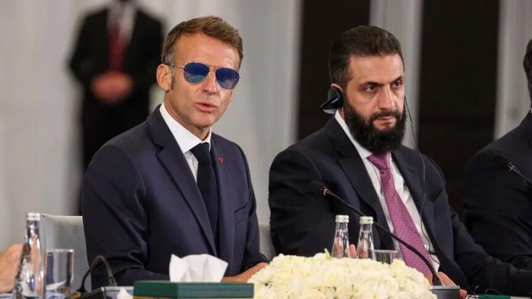 French President Emmanuel Macron wearing sunglasses sitting alongside a Syrian official during a formal meeting.