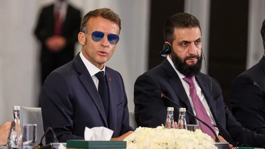 French President Emmanuel Macron wearing sunglasses sitting alongside a Syrian official during a formal meeting.