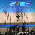 A high-angle view of a lone speaker on a large stage at an international summit, with media cameras in the foreground.
