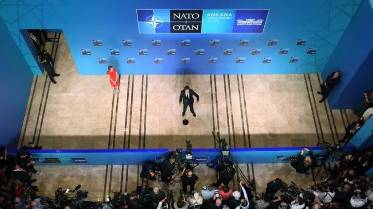 A high-angle view of a lone speaker on a large stage at an international summit, with media cameras in the foreground.