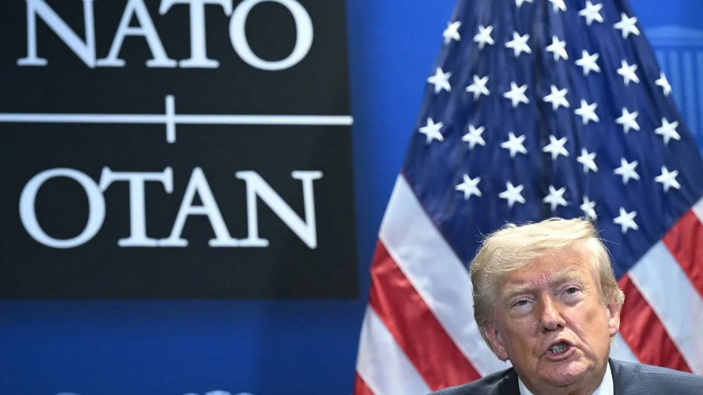 Donald Trump speaking in front of a NATO and US flag.