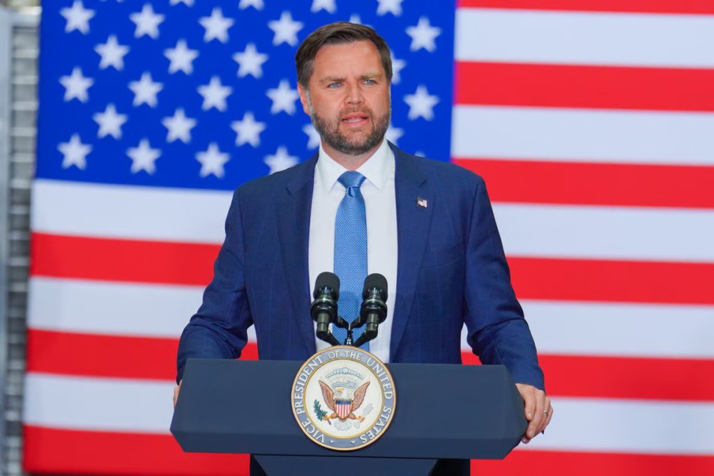 JD Vance speaking at a podium with the Vice President seal, positioned in front of a large American flag.