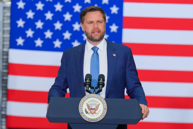 JD Vance speaking at a podium with the Vice President seal, positioned in front of a large American flag.