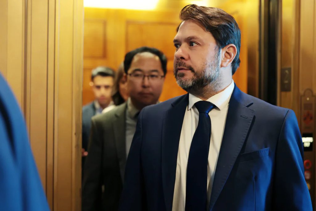A man in a suit and tie walking through an open door, with other professionals following behind him.