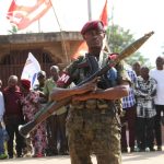 A soldier in camouflage uniform holding a rocket-propelled grenade launcher in a public setting.