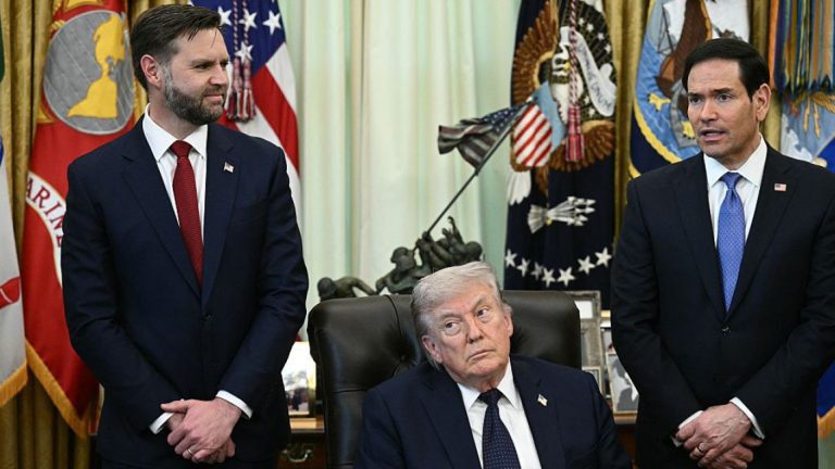 Donald Trump sits at a desk in the Oval Office flanked by two other men in suits.
