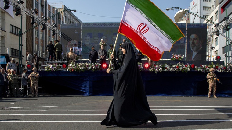 A woman in a black chador holding an Iranian flag during a public event or parade.