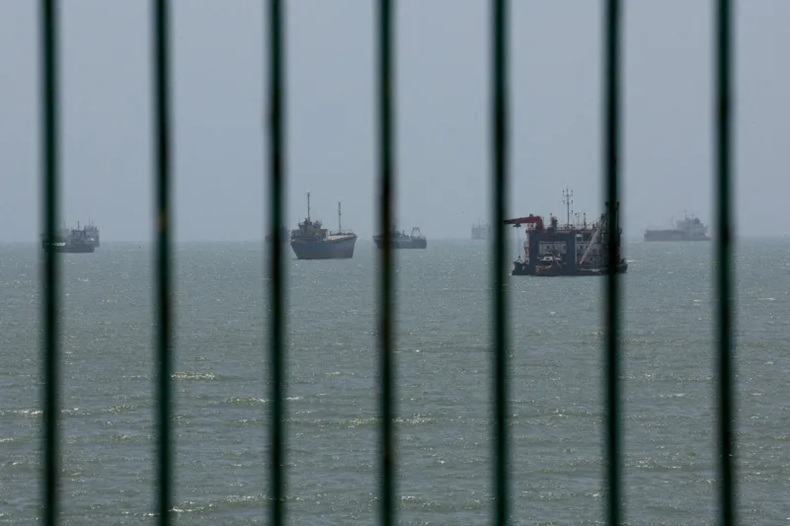 View of cargo ships floating in open water seen through a dark metal fence or grate.