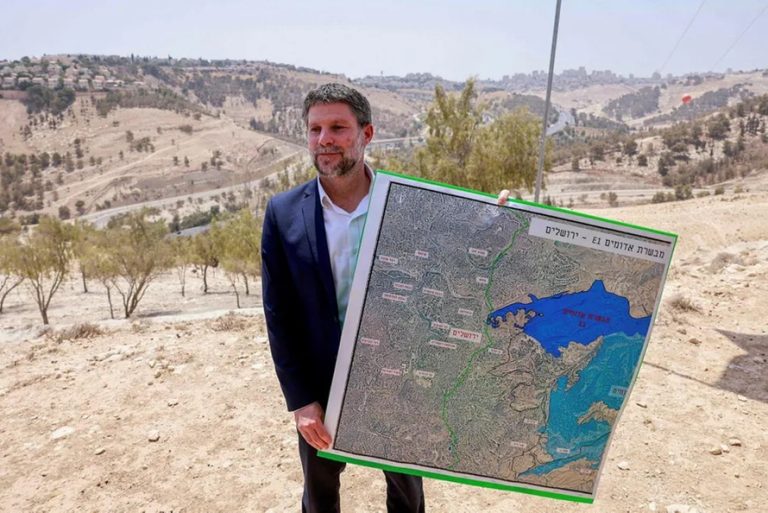 Bezalel Smotrich holding a large map of the E1 area near Jerusalem outdoors.