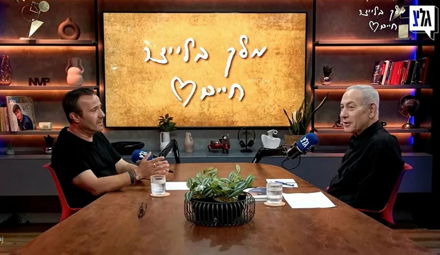 Benjamin Netanyahu sitting at a wooden studio desk during a podcast interview with Hebrew text glowing on a background screen.