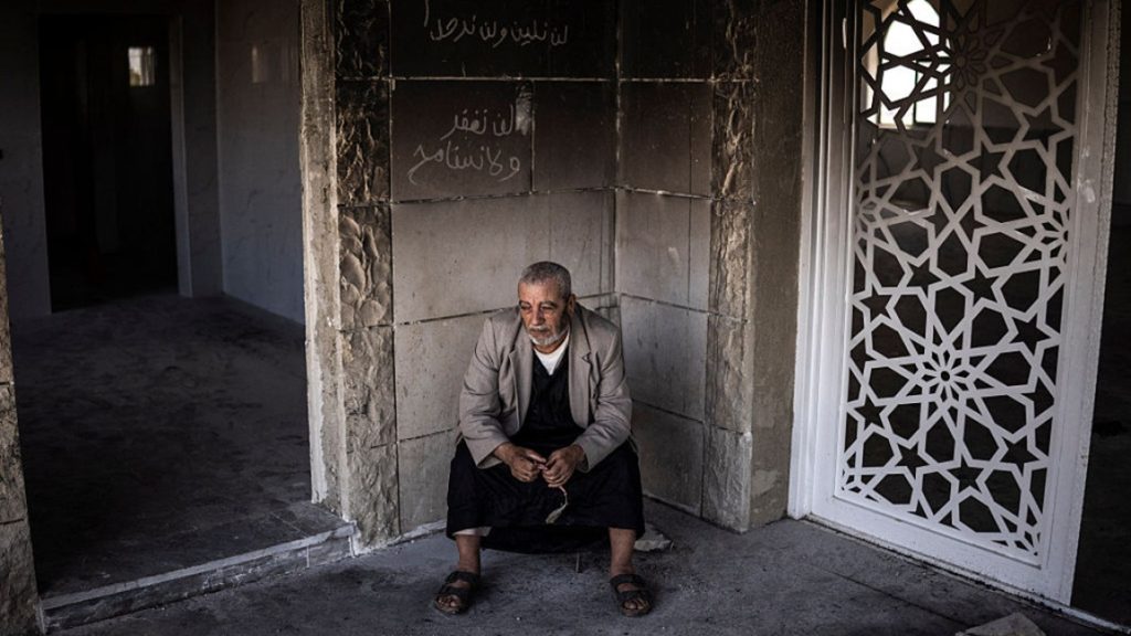 An elderly man sitting alone in a damaged building with Arabic graffiti on the wall.