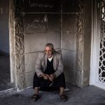 An elderly man sitting alone in a damaged building with Arabic graffiti on the wall.