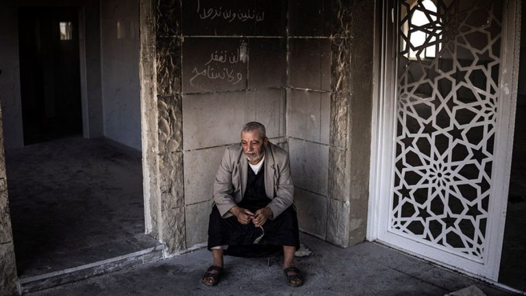 An elderly man sitting alone in a damaged building with Arabic graffiti on the wall.