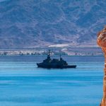 Naval warship sailing in calm blue waters past a steep red rock cliff with mountains in the background.