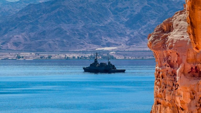 Naval warship sailing in calm blue waters past a steep red rock cliff with mountains in the background.