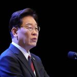 South Korean politician Lee Jae-myung speaking into microphones against a dark background.
