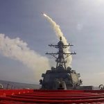 Guided missile launching from the deck of a naval destroyer creating a trail of smoke in the sky.