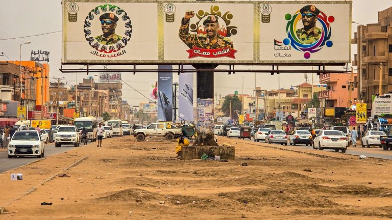 Dusty urban street in Sudan featuring a large billboard displaying military leaders alongside civilian cars.