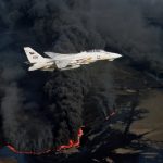 A U.S. Navy fighter jet flying low over burning oil fields with thick black smoke.