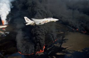 A U.S. Navy fighter jet flying low over burning oil fields with thick black smoke.