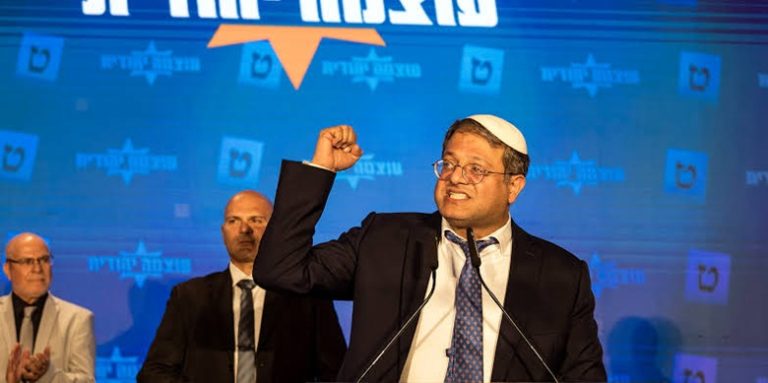 Itamar Ben-Gvir raising his fist while speaking at a podium in front of political banners