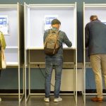 Three voters standing inside private voting booths filling out election ballots.