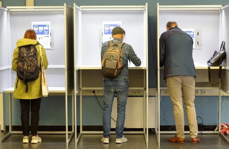 Three voters standing inside private voting booths filling out election ballots.