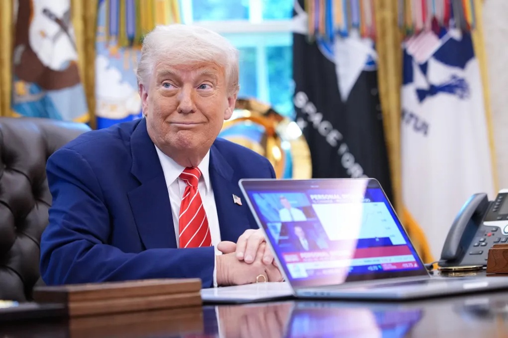 President Donald Trump sitting at a desk with a laptop displaying financial charts in the Oval Office.