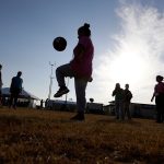 Silhouette of a person juggling a soccer ball outdoors against a bright sunlit sky with onlookers.