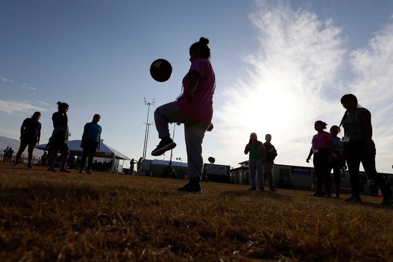 Silhouette of a person juggling a soccer ball outdoors against a bright sunlit sky with onlookers.