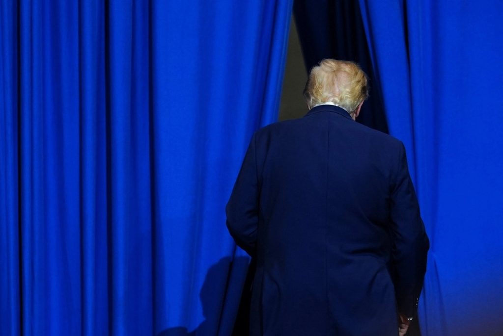 Donald Trump seen from behind walking away through an opening between heavy blue curtains.
