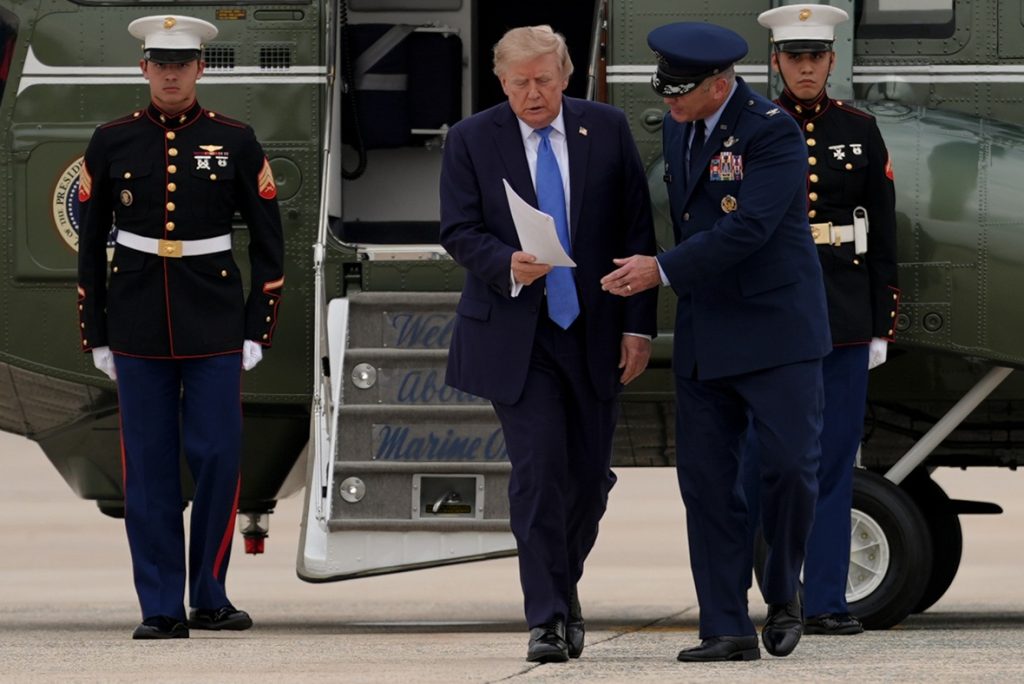 Donald Trump walking out of a Marine One helicopter holding papers, accompanied by military officials.