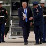 Donald Trump walking out of a Marine One helicopter holding papers, accompanied by military officials.