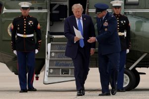 Donald Trump walking out of a Marine One helicopter holding papers, accompanied by military officials.