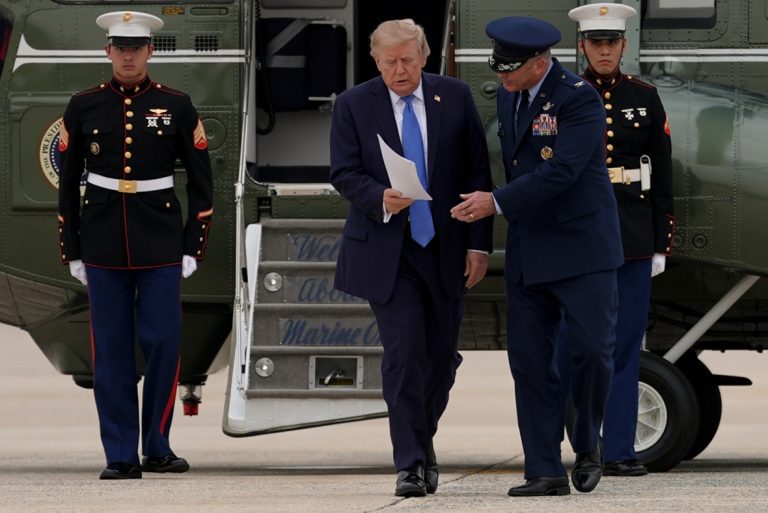 Donald Trump walking out of a Marine One helicopter holding papers, accompanied by military officials.