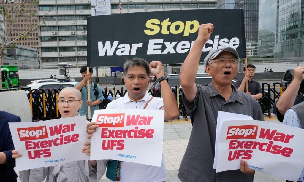 Activists holding protest signs and a banner reading "Stop War Exercise" in front of government buildings.
