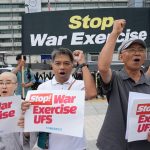 Activists holding protest signs and a banner reading "Stop War Exercise" in front of government buildings.