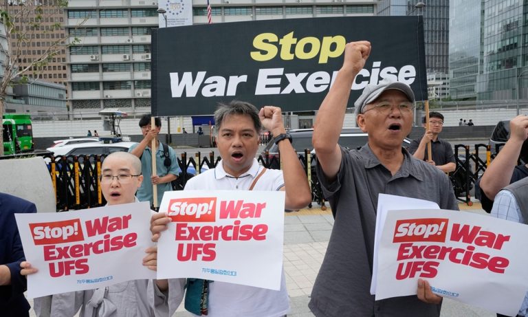 Activists holding protest signs and a banner reading "Stop War Exercise" in front of government buildings.