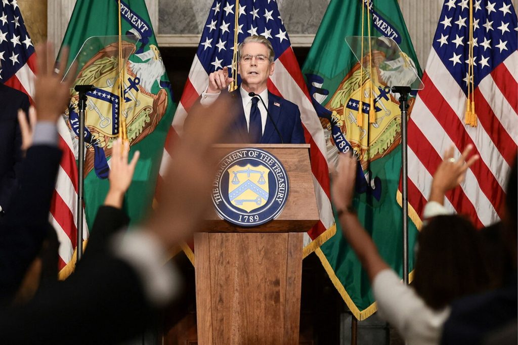 U.S. official speaking at a Department of the Treasury podium surrounded by American flags.