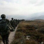 A line of heavily armed soldiers marching across a dry, hilly landscape during patrol operations.