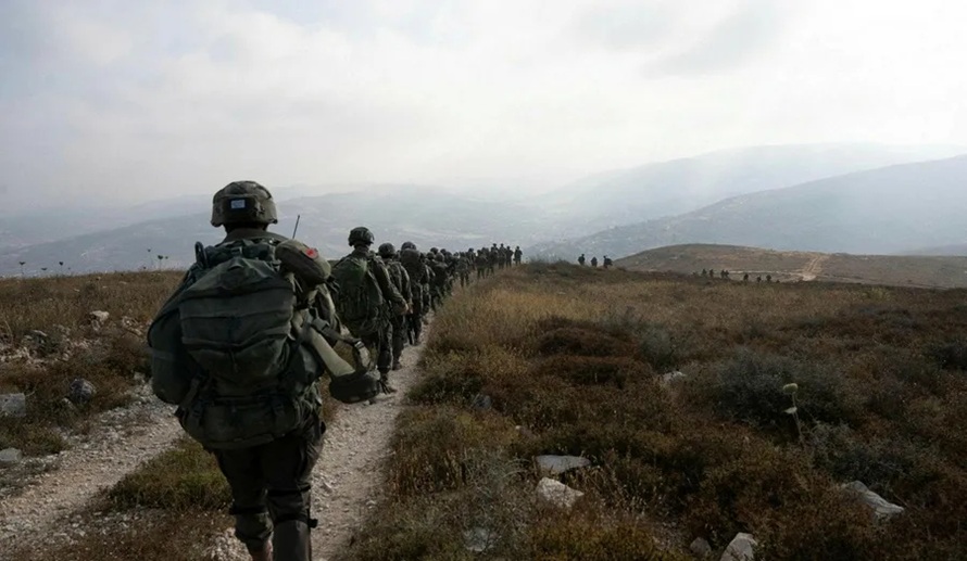 A line of heavily armed soldiers marching across a dry, hilly landscape during patrol operations.